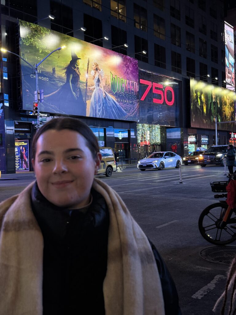 Grayson Donohoe standing to the left of the frame across the street from a Wicked: For Good poster in Times Square